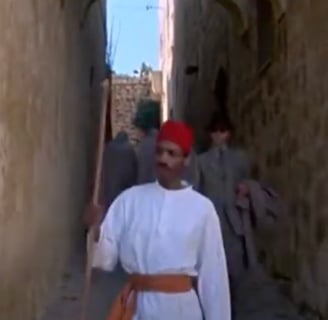 A man in a white robe and a fez walks down a narrow alley.