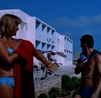 A man and a woman in bathing suits stand in front of a hotel.