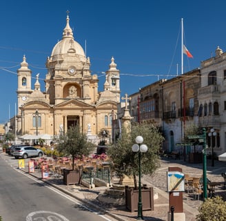 Nadur village square, with restaurant tables and parasols in the sunshine.