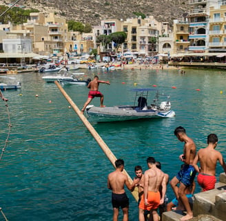 Men and boys in swimming shorts gather on the edge of Xlendi Bay by a greasy pole that juts out over the water.