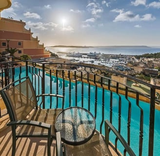 A hotel balcony with two chairs and table, overlooking a swimming pool and Mgarr harbour.