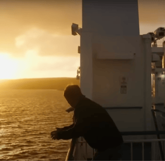 A man leans against the railing of a ship as the sun sets in the distance.