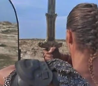 A man in armour and chain mail faces himself in a mirror, atop a cliff.