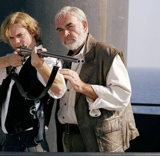 A man holding a gun in front of a submarine conning tower, while another man tutors him.