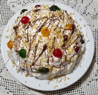 A dome shaped cake covered in whipped cream, multi-coloured glace cherries, chopped nuts and drizzled chocolate. 