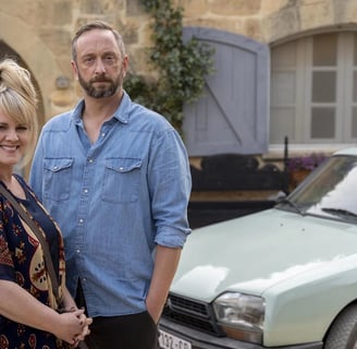 Jean Whiite and Dom from The Madame Blanc Mysteries, standing in front of a tradtional cottage next to a car. 