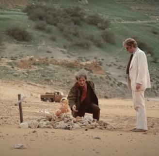 Two men stand on a beach by a grave.