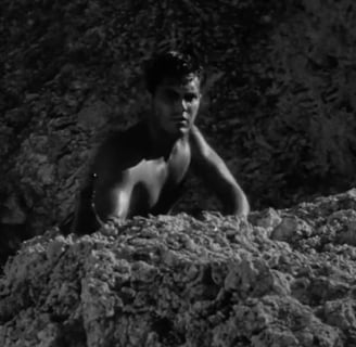 A bare-chested man hides behind a rock.