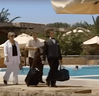 A man and woman are being escorted past a hotel pool a porter, who is carrying their luggage.