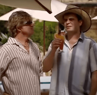 Two men stand underneath a sun parasol  in front of a hotel pool.