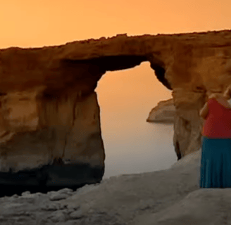 A man and woman stand looking at the sunset through the Azure Window. He has his arm around her shoulder.