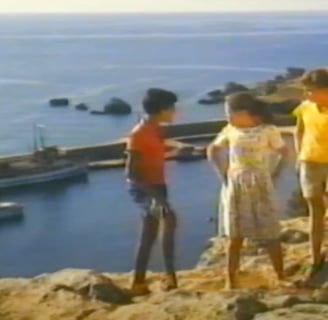 Three children standing on a cliff overlooking a harbour. 