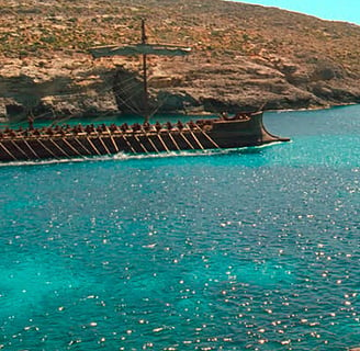 A man dressed as an ancient Greek stands on a cliff in front of a body or water with a galley.