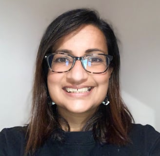 Photo of a brown lady with long dark hair, wearing glasses and a dark top, smiling against a light b