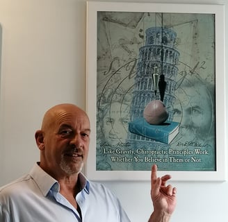Adam Newmanin a blue shirt is standing in front of a chiropractic poster