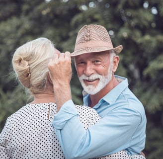 An older man in a fedora and white beard and a stylish older woman are embracing