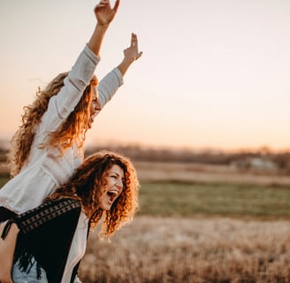 two joyful women with curly long  hair are celebrating, one lifting the other up on her shoulders 