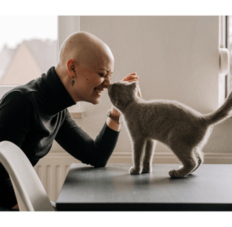 a smiling woman in a black turtle neck and a  bald head and a grey cat are touching nose to nose