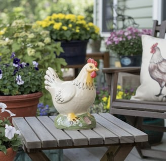 A front porch styled with a  small wooden table holding a ceramic chicken figurine, surrounded by fl