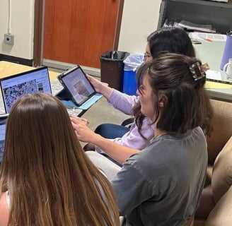 Three students sitting at a table, each with a computer or tablet in front of them reviewing video