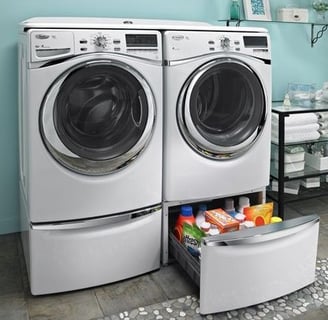 Front-loading washer and dryer in a laundry room with a storage drawer containing detergent and cleaning supplies underneath.