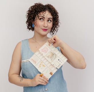 Uma mulher de vestido azul e cabelo cacheado segurando uma paleta de aquarela