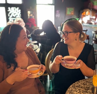 Two women are drinking tea and look at each other smiling. They both are wearing cute little hats.