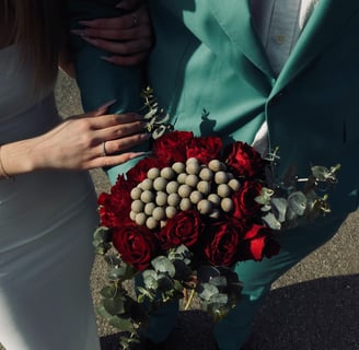 Buchet cu trandafiri roșii, craspedia și eucalipt, ținut de un cuplu.