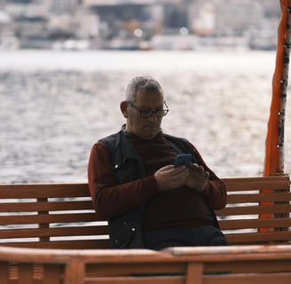 Homem sentado em um pier, camisa bordô e jaqueta verde, de óculos, olhando para o celular 