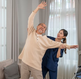 An elderly man and a younger female physical therapist in a living room doing shoulder exercises.