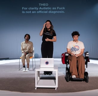 A black person, a white BSL signer, and a mixed-race electric wheelchair user sit on the stage