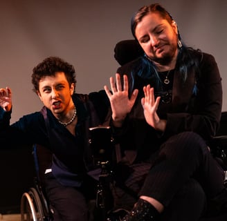 A mixed race manual wheelchair user and a white electric wheelchair user hold up their hands