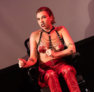 A white person in an electric wheelchair wearing red leather trousers, harness, and tape