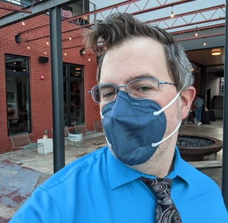 Photo of a 40ish white person with graying blond hair, glasses, a blue respirator, & a red paisley tie, standing on a patio.