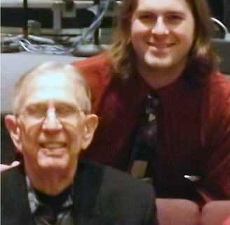 Photograph: a 30-ish white person with shoulder-length blond hair and a tie leans over a squinting, 90-ish man in a dark suit