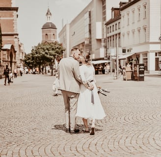 Paar in eleganter Kleidung steht auf einem gepflasterten Platz, im Hintergrund eine Kirche.