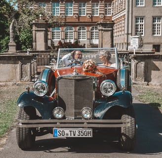 Ein retro Auto mit zwei Personen sitzt davor, lächelnd in der Sonne.
