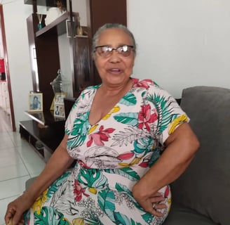 Helena Almeida sentada em casa, vestida com roupa florida, sorrindo para a câmera