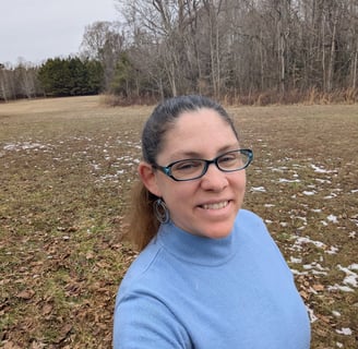 Photo of me, a white woman with long brown hair in a ponytail, wearing a blue shirt & glasses. Field and trees in background