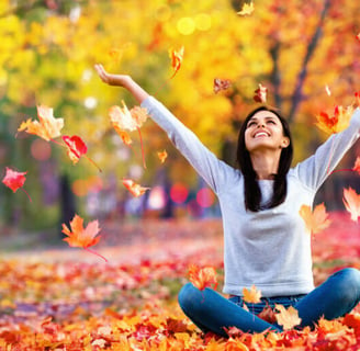 a woman joyfully sitting on a leafy field with her arms up.