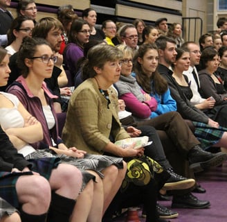 Crowd of people sitting on bleachers, some in kilts, intently watching something
