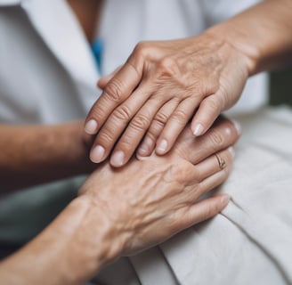 An elderly hand gently rests on top of another, creating a sense of care and companionship. The nails are neatly manicured with a pale color. The background is softly blurred, suggesting an intimate and personal setting.