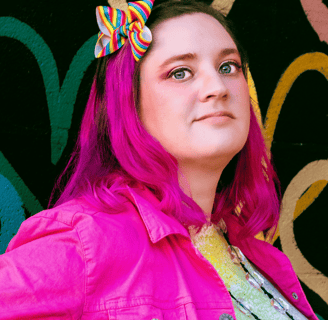 Erika Gritton stands in front of a wall of painted hearts. Her hair is pink with a rainbow bow in it