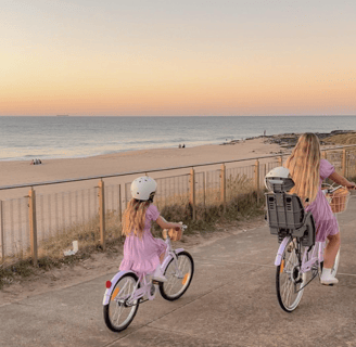 Mère et fille à vélo en bord de mer au coucher du soleil, toutes deux en robe violette.