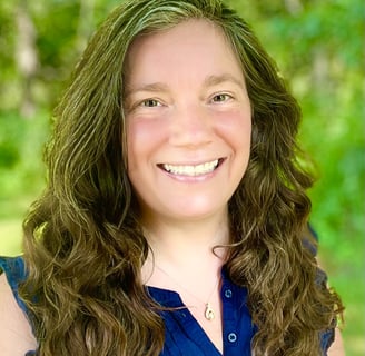 Shannon Gale, with long brunette hair, and a navy shirt, smiling in front of a green woodland scene