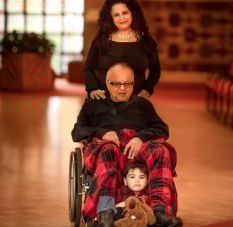 A woman pushes a frail man in a wheelchair, a young child holds a bear at the man's feet.
