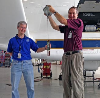Two persons holding a dropsonde and its parachute beside Global Hawk drone for science and weather.