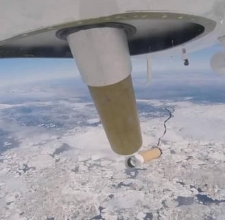 Under belly of plane image of dropsonde departing aircraft above a snow covered landscape far below.