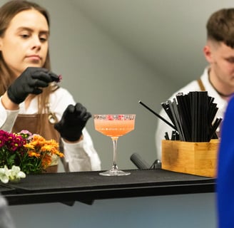 Baro darbuotoja su juodomis pirštinėmis dekoruoja elegantišką kokteilį rožiniu gėrimu taurėje. Priek