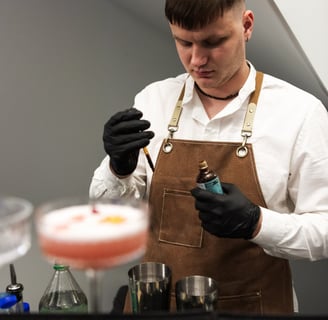 Barmenas, dėvintis baltus marškinius ir rudos spalvos prijuostę, ruošia kokteilį profesionalioje bar